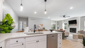 Une cuisine moderne avec des meubles blancs, un lave-vaisselle et un évier. On y trouve également un coin repas avec un canapé et un téléviseur fixé au mur.