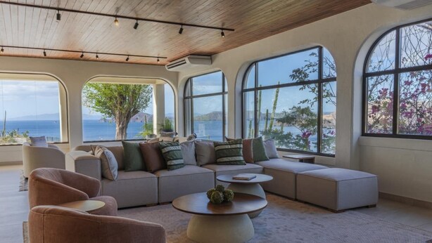 Uma sala de estar ampla com um sofá seccional grande, uma mesa de centro redonda e várias janelas com vista para o mar e vegetação.