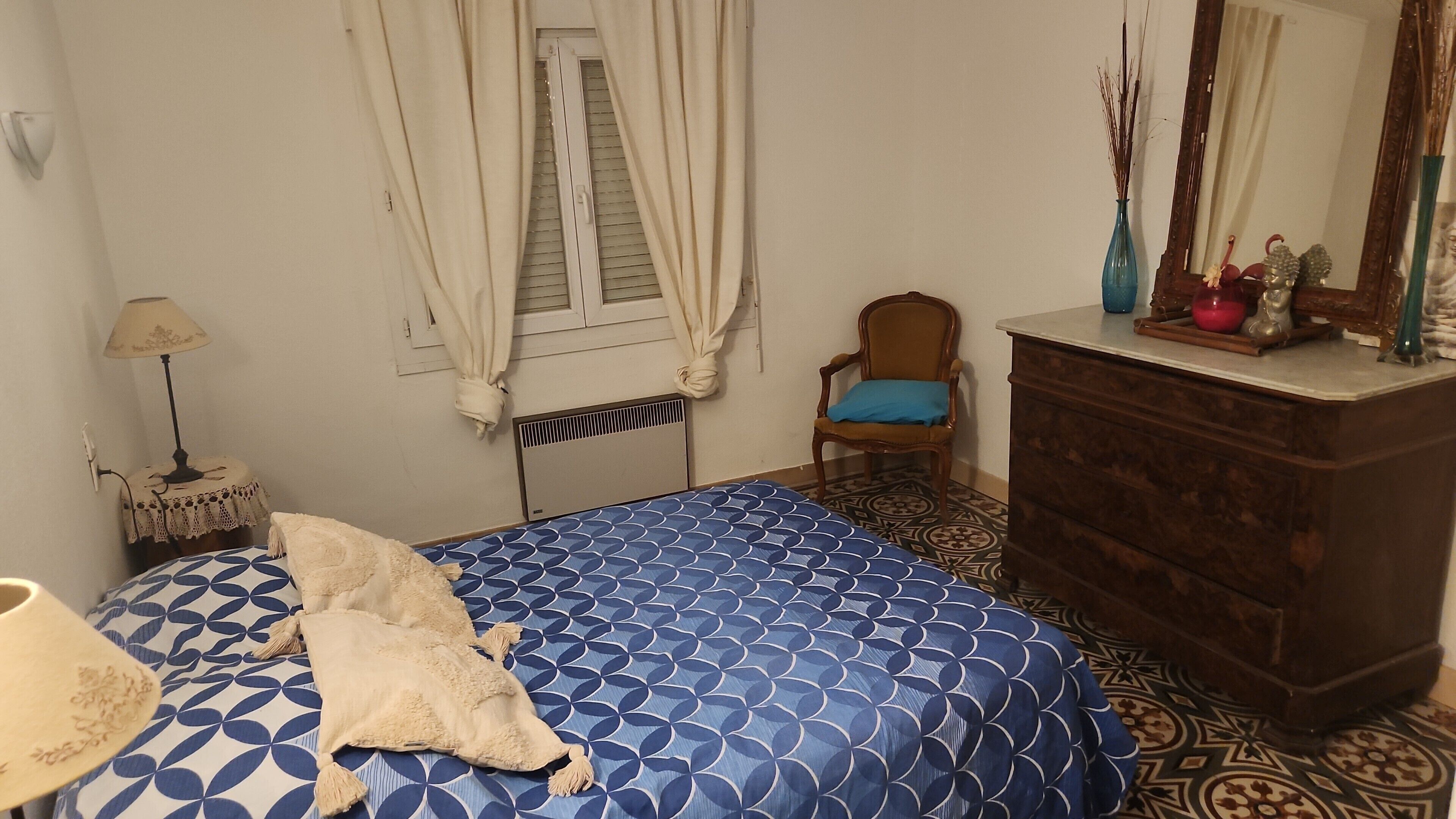 Ein Schlafzimmer mit Bett, Stuhl, Kommode, Lampe und einem Fenster mit Vorhängen.