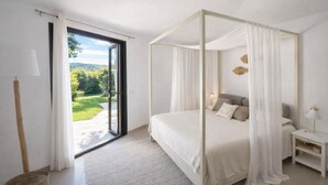 Un dormitorio con cama con dosel, una mesita de noche, una lámpara y vistas a un jardín a través de una puerta corredera.
