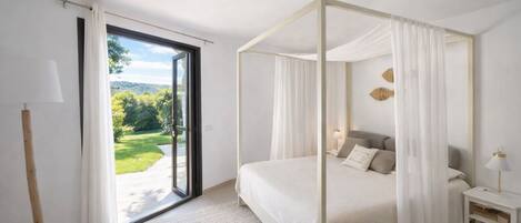 A bedroom with a four-poster bed, a nightstand, a lamp, and a view of a garden through a sliding door.