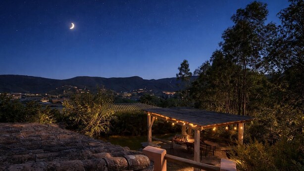 Un patio exterior iluminado por la noche, con pérgola, luces colgantes y vistas a colinas distantes y una luna creciente.