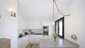 Una cocina moderna con armarios blancos, electrodomésticos de acero inoxidable, una lámpara colgante y vistas a través de una puerta.