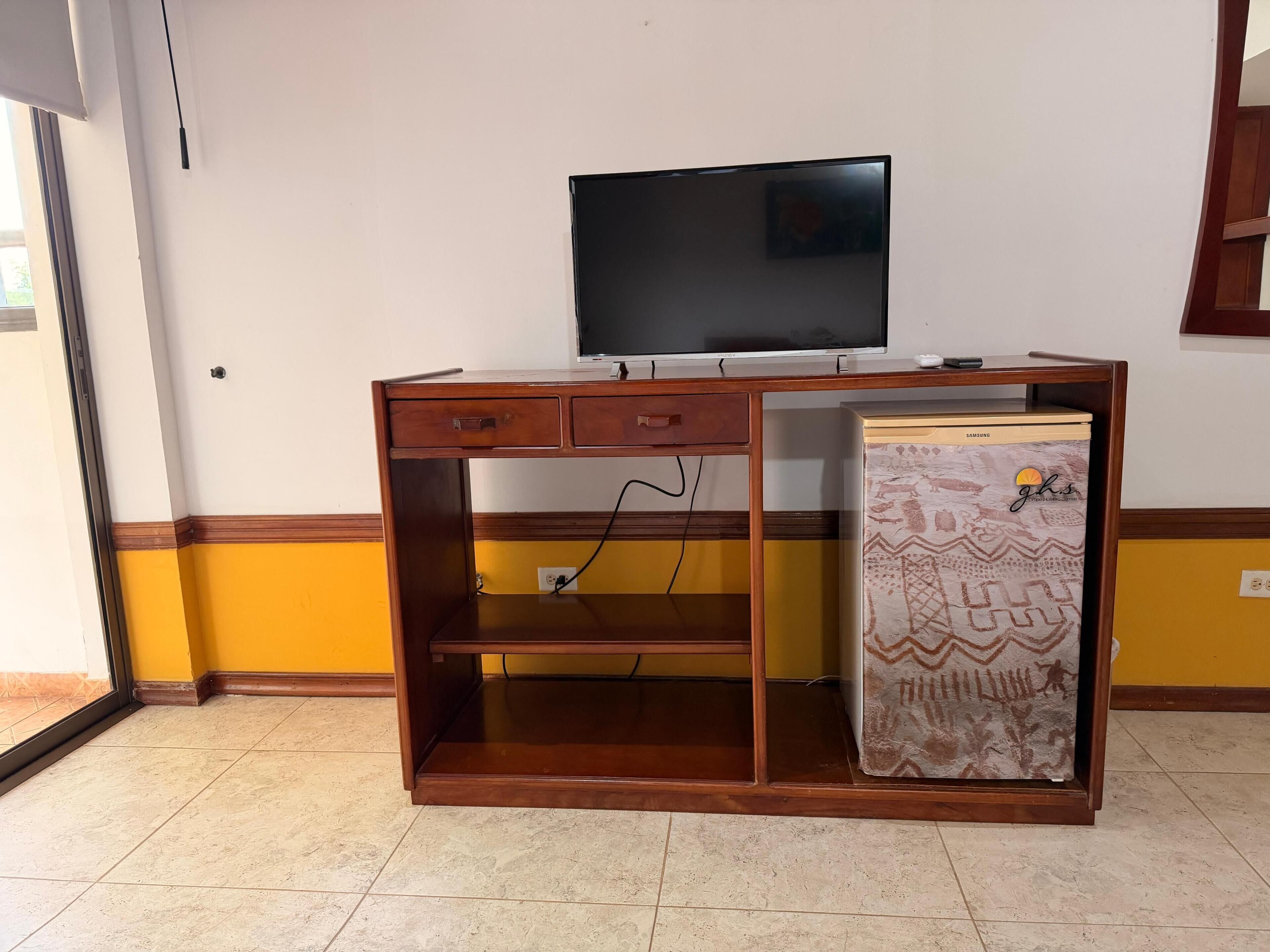 A wooden desk with a television, a patterned refrigerator, and a tiled floor.