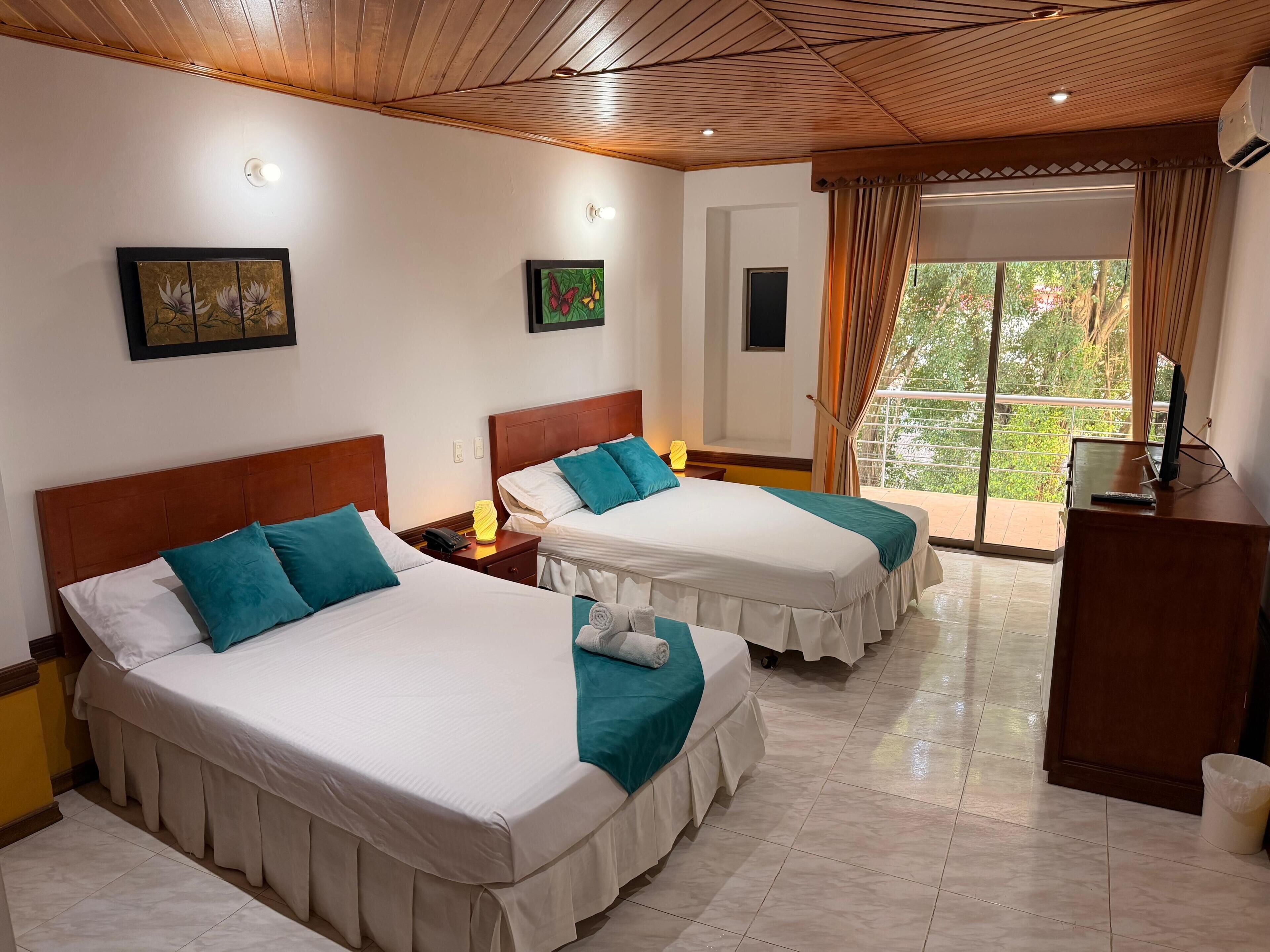 Habitación de hotel con dos camas, televisión, escritorio y balcón con vistas.