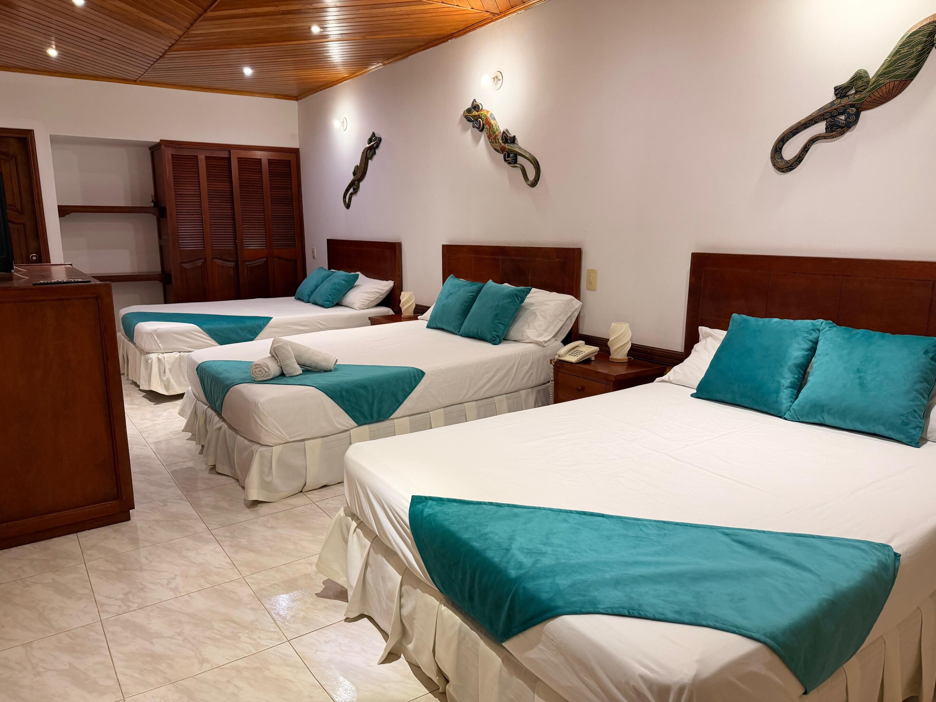 A hotel room with three beds, each with teal bedding and white pillows, wooden headboards, and a wooden wardrobe.
