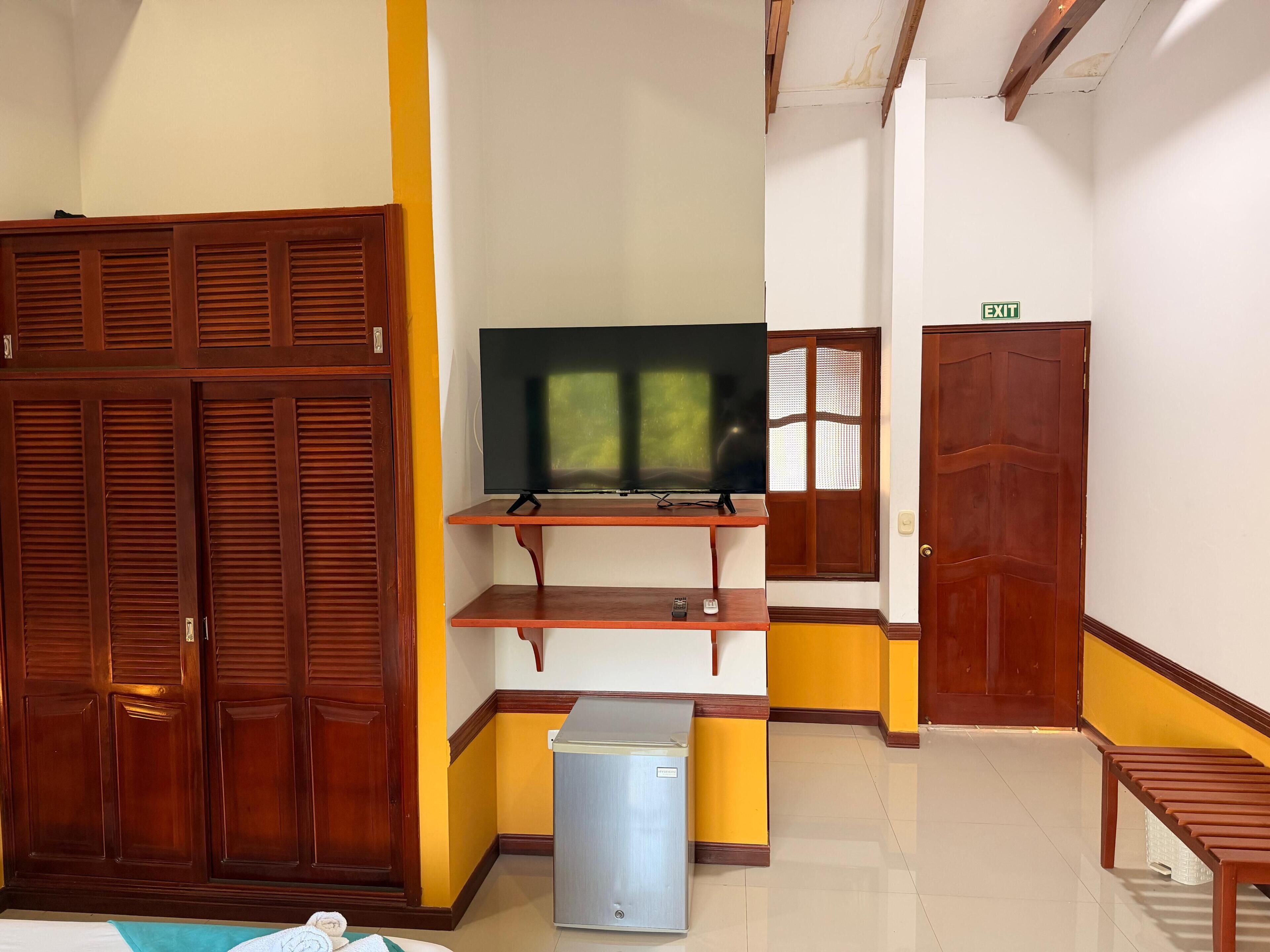 A room with a wooden wardrobe, a television on a wooden stand, a small refrigerator, and a bench.