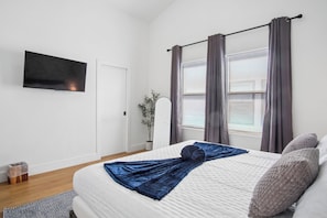 Ein Schlafzimmer mit Bett, Fernseher, einem Fenster mit Vorhängen, einer Zimmerpflanze und einer Tür.