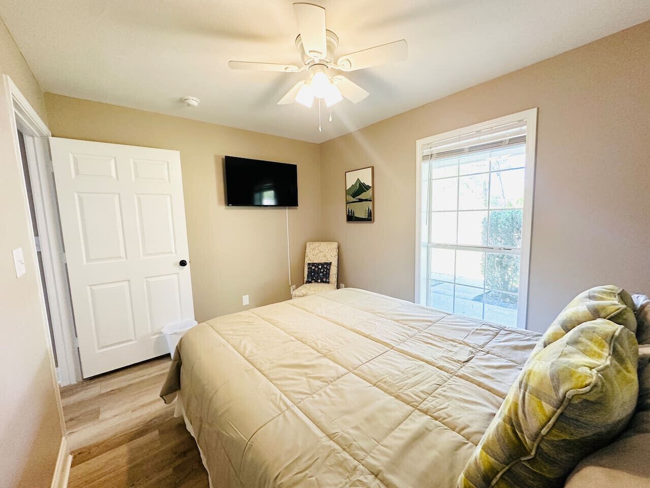 A bedroom with a bed, a ceiling fan, a TV, a window, and a door.