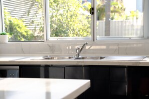 A kitchen with a double sink, white marble countertop, and a window with a view of greenery. - The Beachside Gathering (Anna Bay)