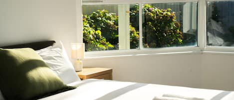 A neatly made bed with a pillow, a bedside table with a lamp, and a window with a view of greenery.