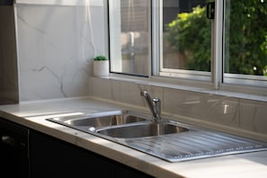 A kitchen with two stainless steel sinks, a potted plant, and a window with a view of greenery. - The Beachside Gathering (Anna Bay)