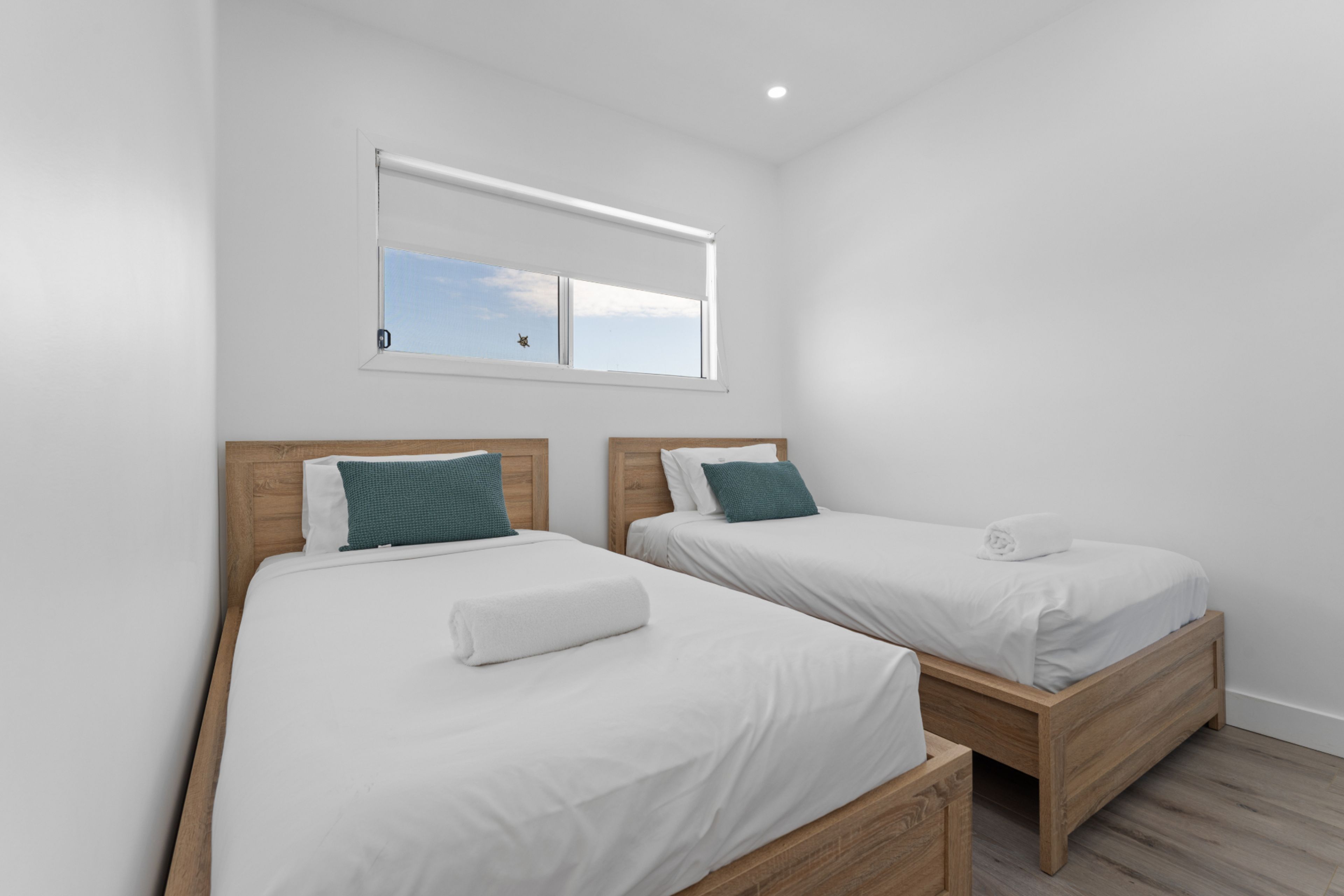 Ein Zimmer mit zwei Einzelbetten, jeweils mit einem hölzernen Kopfteil, weißer Bettwäsche und einem türkisfarbenen Kissen. Über den Betten befindet sich ein Fenster mit Blick auf den Himmel.