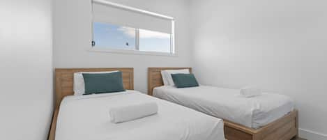 A room with two single beds, each with a wooden headboard, white bedding, and a teal pillow. There is a window above the beds with a view of the sky.