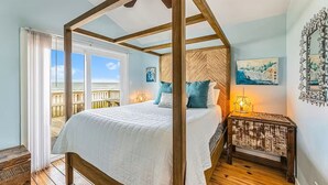 Un lit à baldaquin donnant sur la plage, un plancher en bois et une table de chevet avec une lampe.