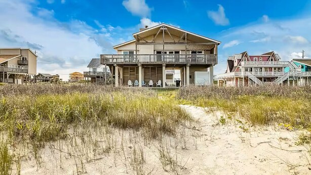 Une maison située en bord de mer, dotée d’un balcon, d’une véranda avec des chaises et d’un chemin de sable menant à l’entrée.