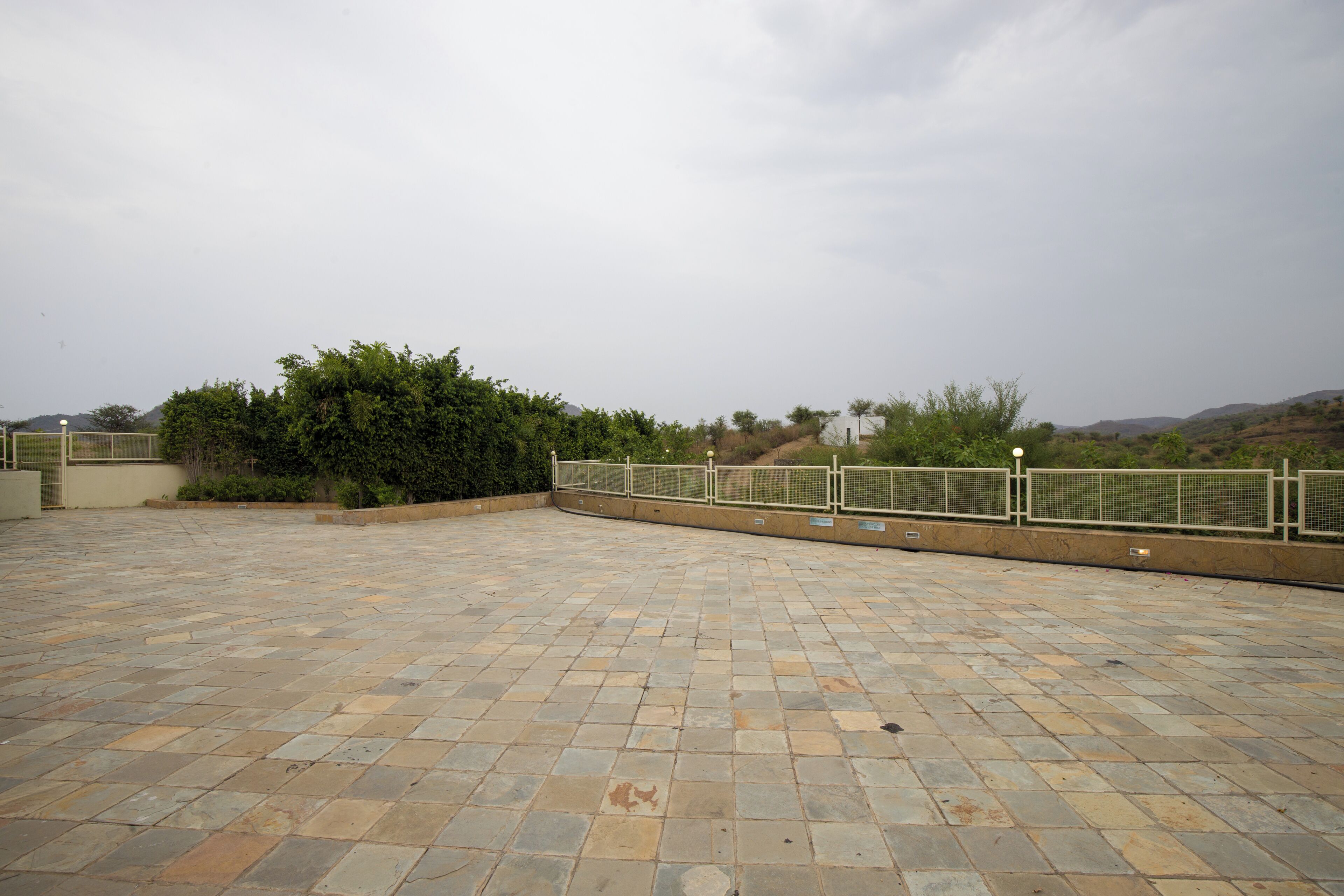 Una terraza pavimentada con barandilla de metal, rodeada de vegetación y colinas al fondo.