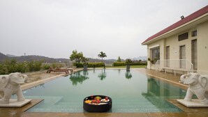 Una piscina con un jacuzzi en el centro, rodeada por dos estatuas de elefantes, un edificio con techo rojo y un paisaje montañoso al fondo.