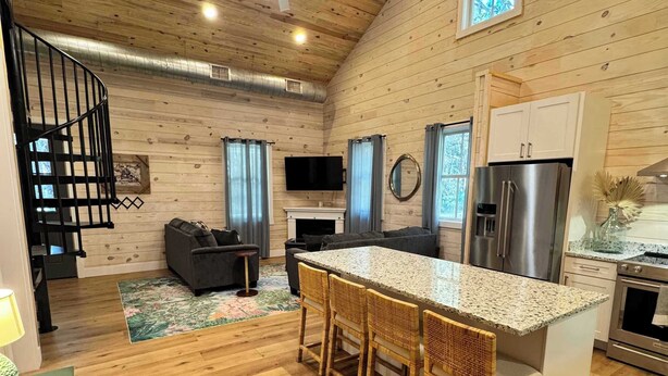 A spacious living area with a wooden ceiling, a central island with bar stools, a fireplace, and a kitchen with modern appliances. - Carvin Cottage - Cozy 2bdr with Loft- 4 Passenger Golf Cart Included (Daufuskie Island)