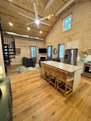 Een ruime woonruimte met een houten plafond, een centraal eiland met barkrukken, een open haard en een keuken met moderne apparatuur.