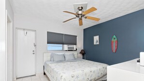 Un dormitorio con cama, ventilador de techo, ventana con persianas, aire acondicionado instalado en la pared y decoración con una tabla de surf.