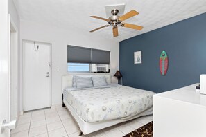 Une chambre avec un lit, un ventilateur de plafond, une fenêtre avec des stores, un climatiseur fixé au mur et une décoration sur le thème du surf.