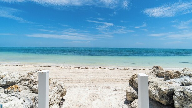 Uma praia com céu claro, mar calmo e um primeiro plano rochoso.