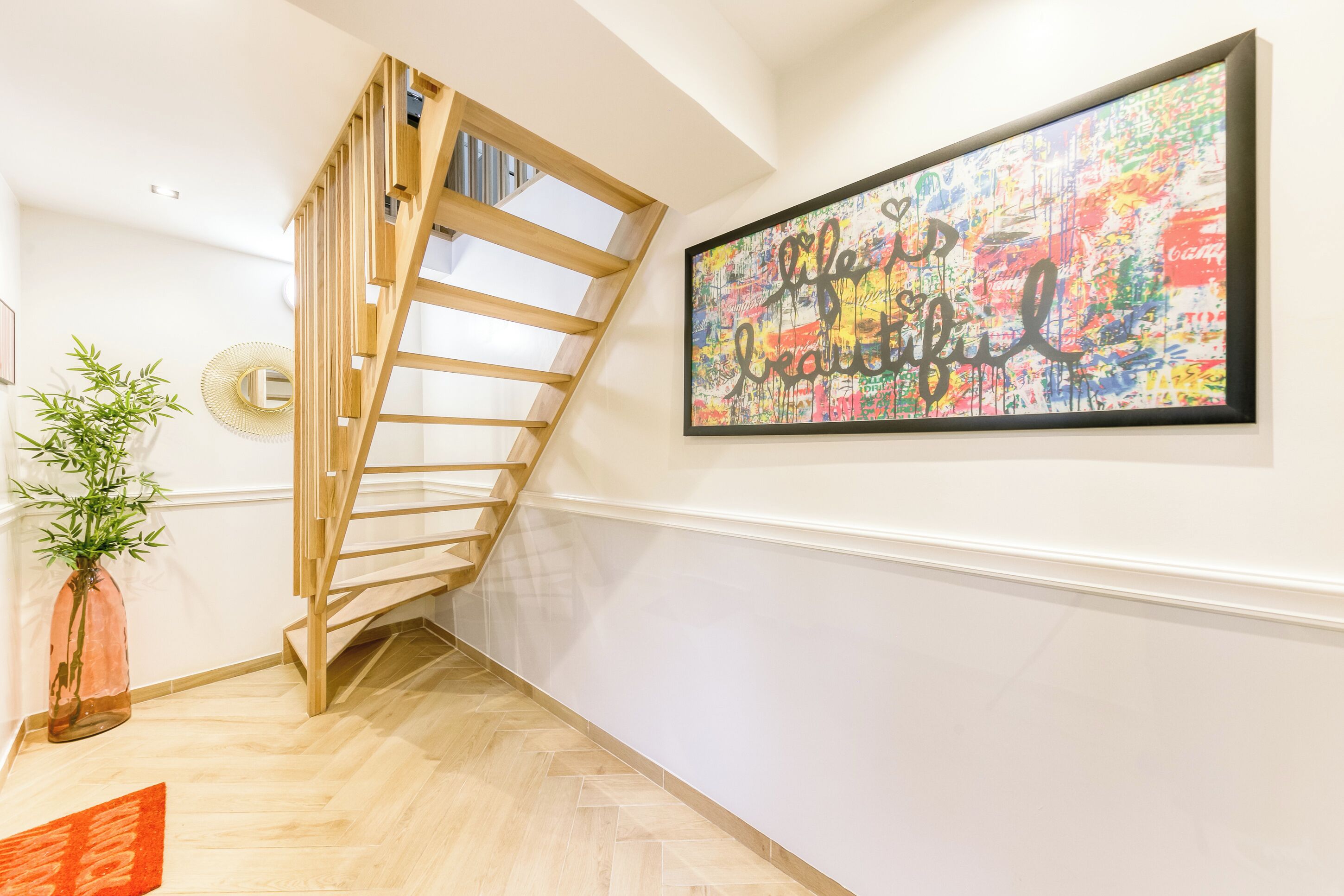 Eine Treppe mit hölzernem Geländer, ein an der Wand befestigtes abstraktes Gemälde, ein Blumentopf und ein kleiner Tisch mit einer Vase.