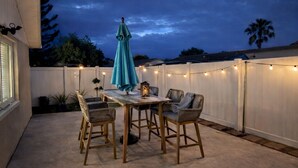 Un patio al aire libre con una mesa, sillas, luces colgantes y un paraguas azul.