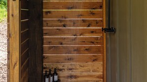 A wooden door with a small table holding bottles, a showerhead above, and a concrete floor. - Cabin w/ Sauna + Hot Tub - Luxury Woods Retreat (Logan)