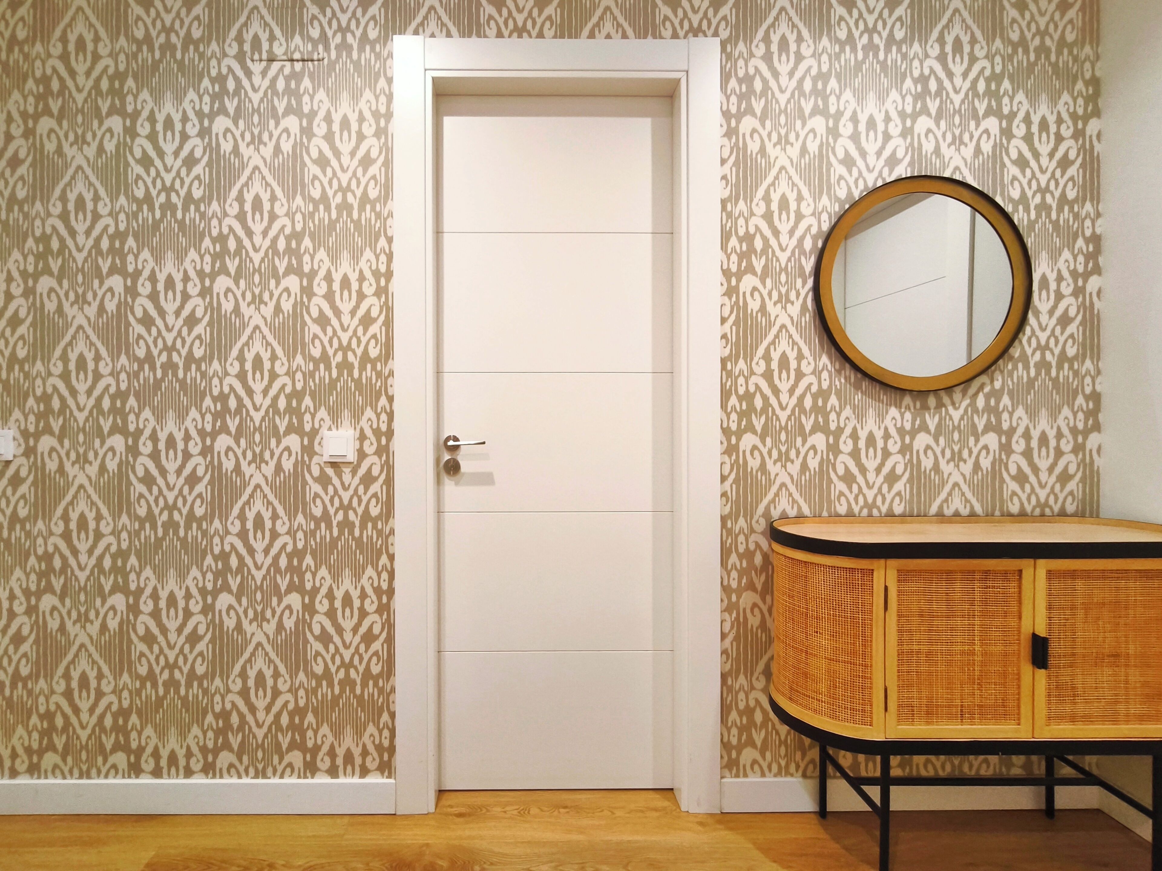 A room with patterned wallpaper, a white door, a round mirror, and a wicker cabinet.