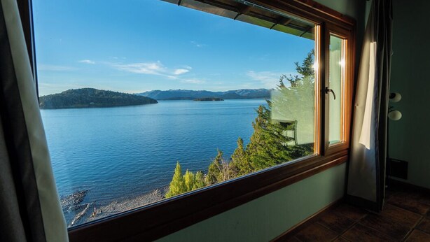 A room with a large window overlooking a lake, a door, and a chair near the window. - Casa Siete - Your home in Bariloche (San Carlos de Bariloche)
