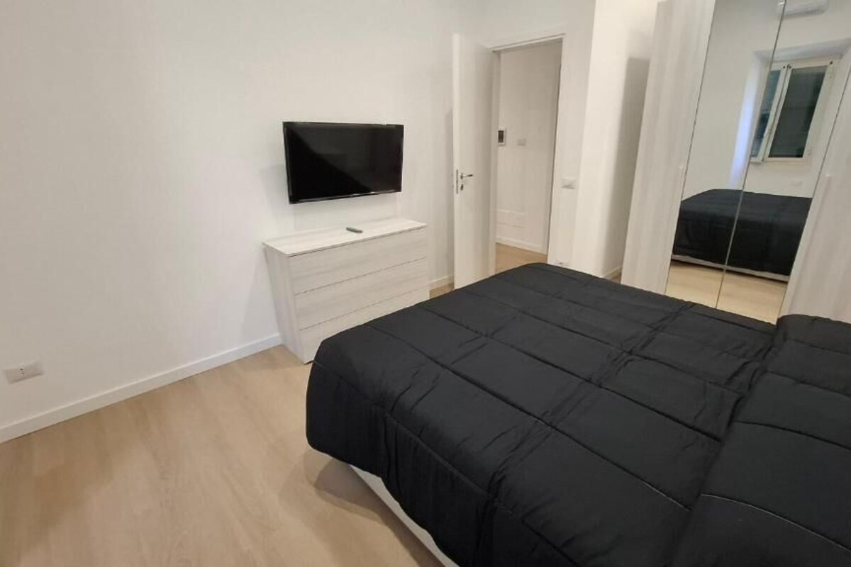 A bedroom with a bed, a TV on a stand, a door, and a mirror reflecting the room.