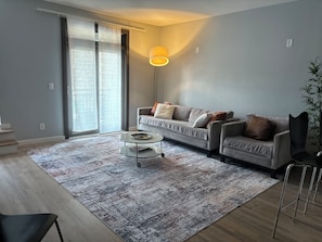 Een moderne woonkamer met een grijze bank, een salontafel, een groot raam met zonwering en een vloerlamp.