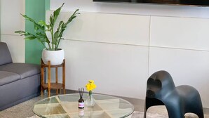 A living room with a round glass coffee table, a black elephant-shaped stool, a potted plant, and a wall-mounted TV. - Room-charming B&B Retreat in Vibrant Phr (Bangkok)