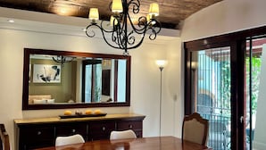 Uma sala de jantar com uma mesa de madeira, cadeiras brancas, um lustre, um espelho grande e vista através de uma porta de vidro.