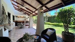 Uma varanda coberta com uma mesa de jantar, cadeiras, uma cadeira de balanço de macramé, um vaso de planta e vista para um jardim.