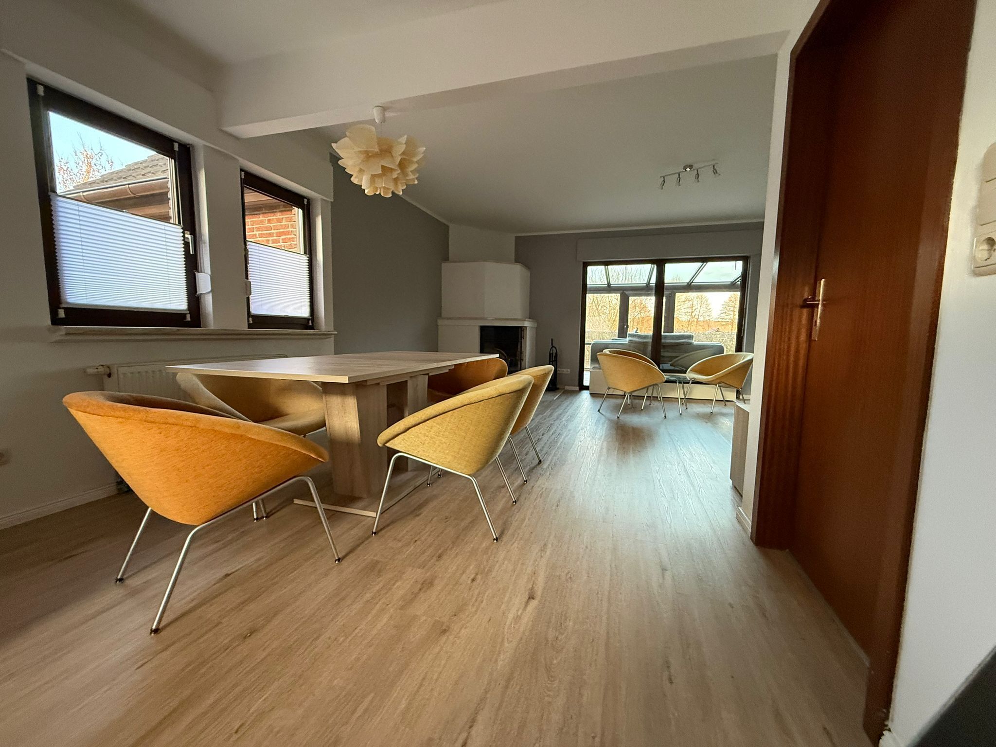 Ein modernes Interieur mit einem Essbereich, der einen Holztisch und gelbe Stühle umfasst, einer Küche mit weißen Schränken und großen Fenstern, die viel natürliches Licht hereinlassen.
