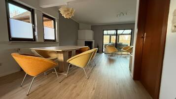 Ein modernes Interieur mit einem Essbereich, der einen Holztisch und gelbe Stühle umfasst, einer Küche mit weißen Schränken und großen Fenstern, die viel natürliches Licht hereinlassen.