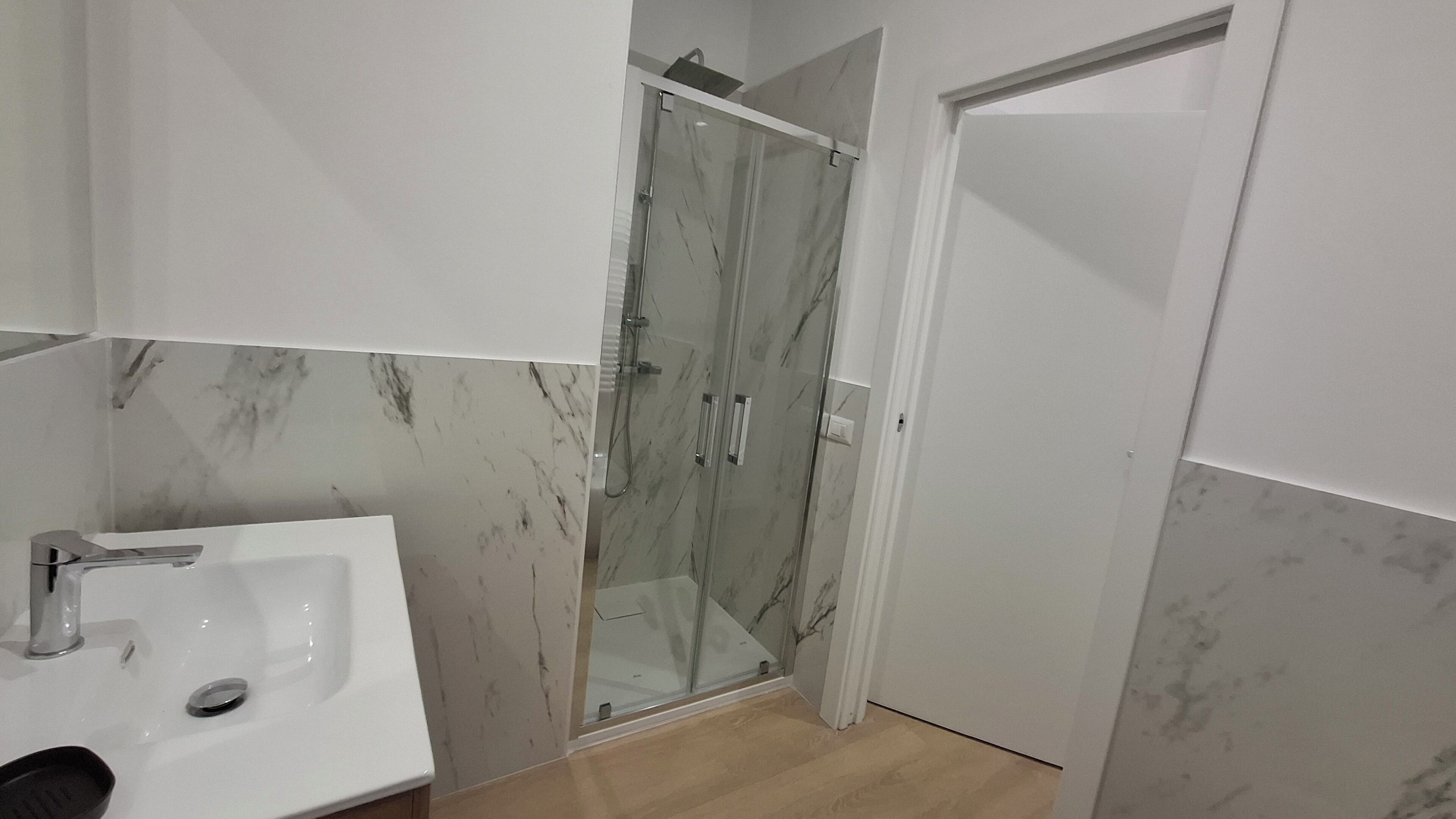 Un bany amb dutxa de vidre, un lavabo blanc i parets amb dibuixos de marbre.