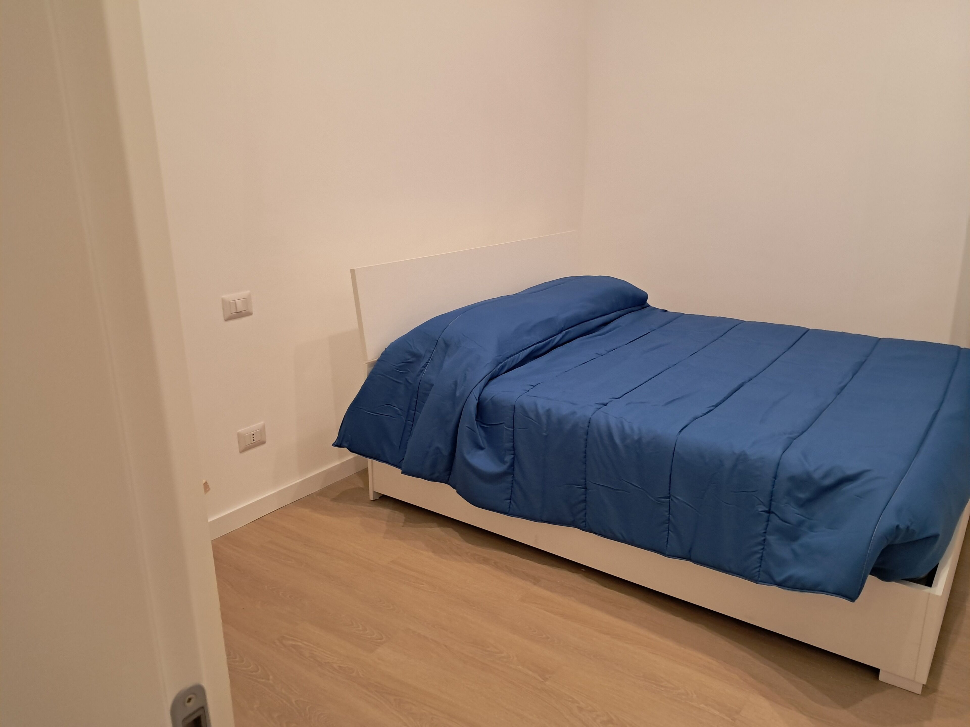 A single bed with a white frame and a blue bedspread in a room with white walls and wooden flooring.