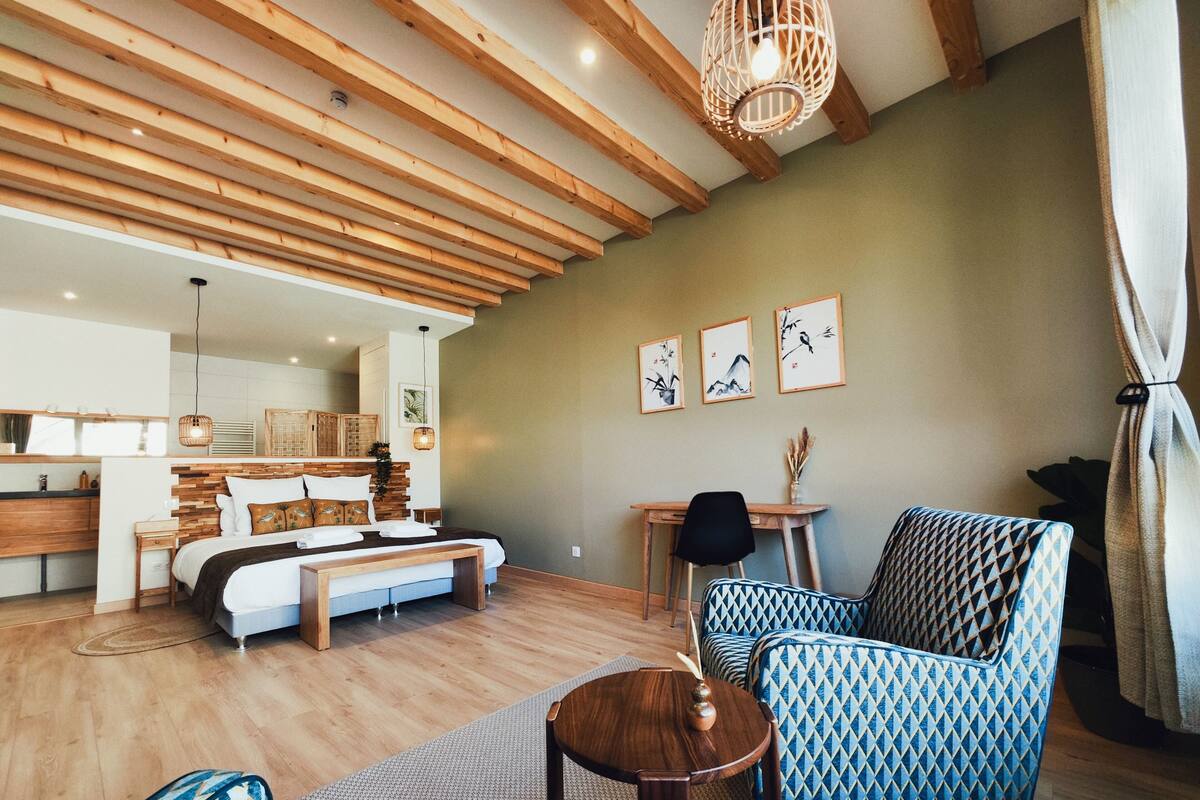A spacious room with a wooden ceiling, a bed with a wooden headboard, a desk, and patterned chairs.