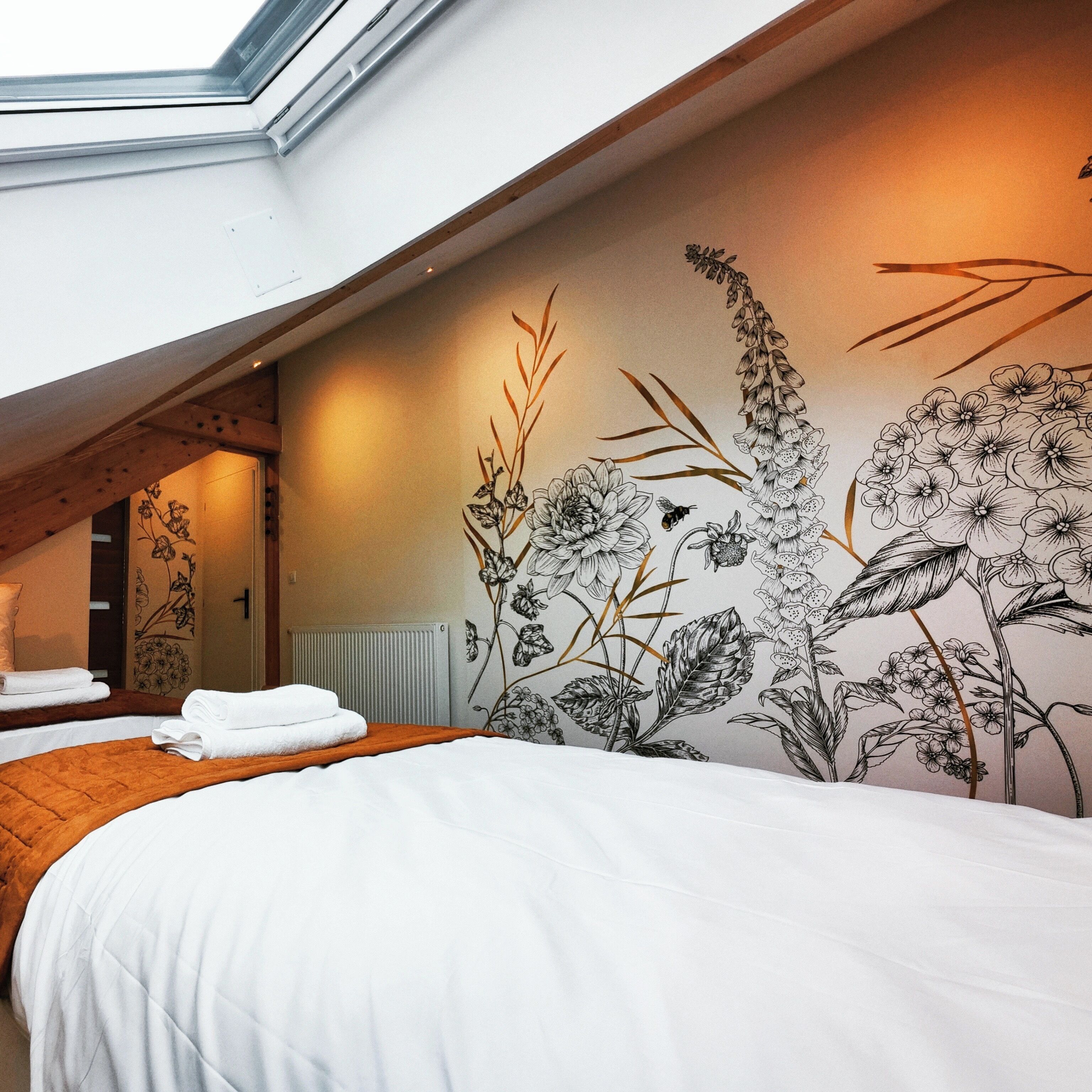 Un dormitorio con tragaluz, una cama con ropa blanca y un mural floral con abejas.