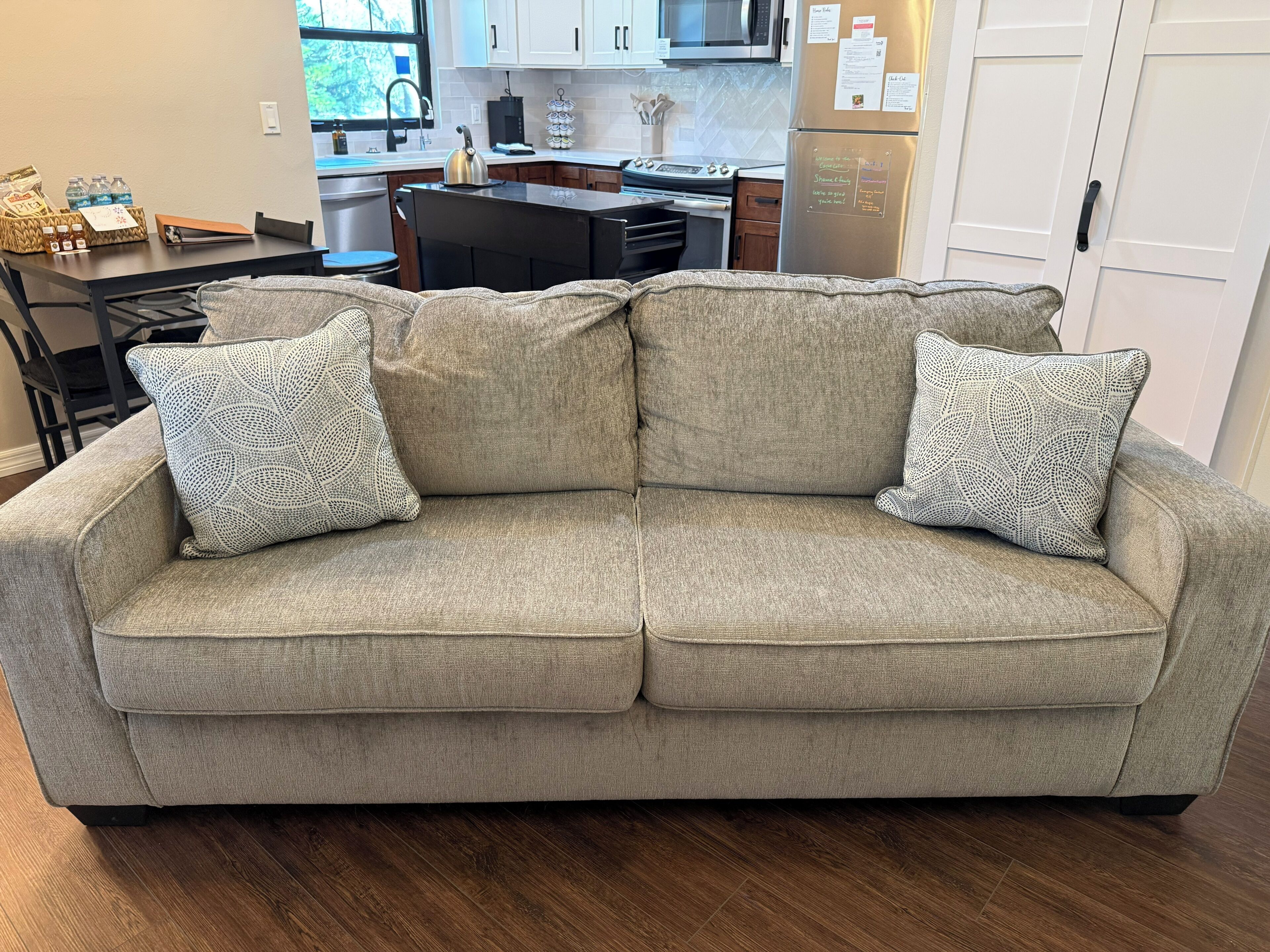 A grey sofa with patterned cushions in a modern kitchen with white cabinets and a stainless steel refrigerator.