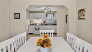 Uma área de refeições com uma mesa e cadeiras brancas, um ventilador de teto e uma cozinha ao fundo.
