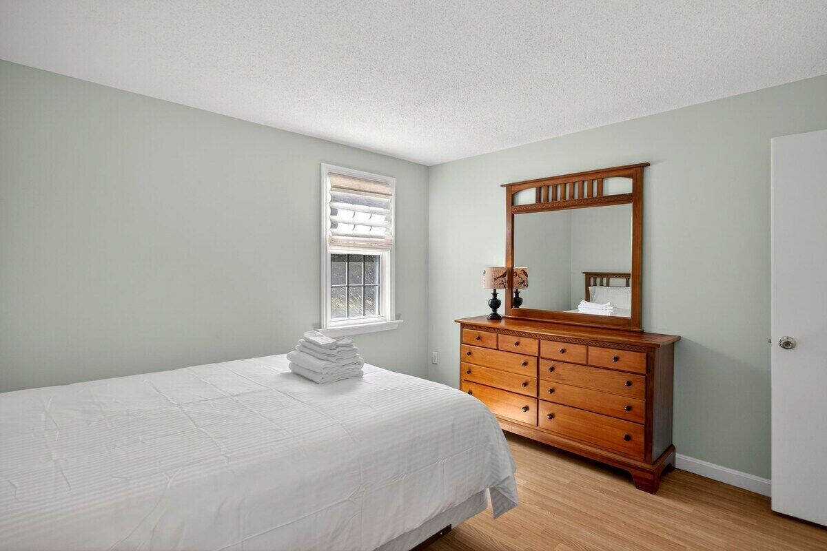 A bedroom with a bed, dresser, mirror, window with blinds, and a door.
