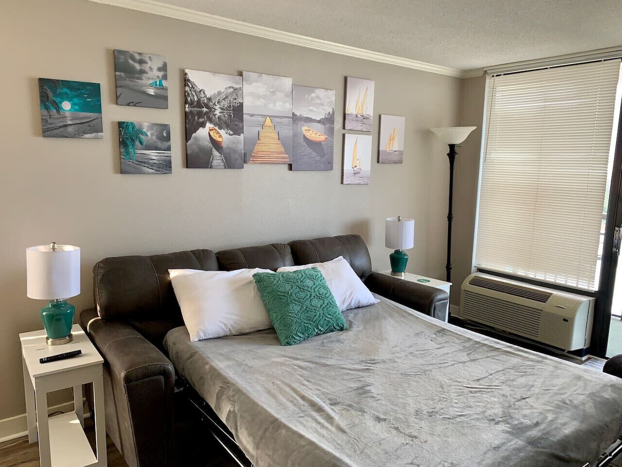 A hotel room with a bed, two bedside tables, a lamp, a heater, and a window with blinds.