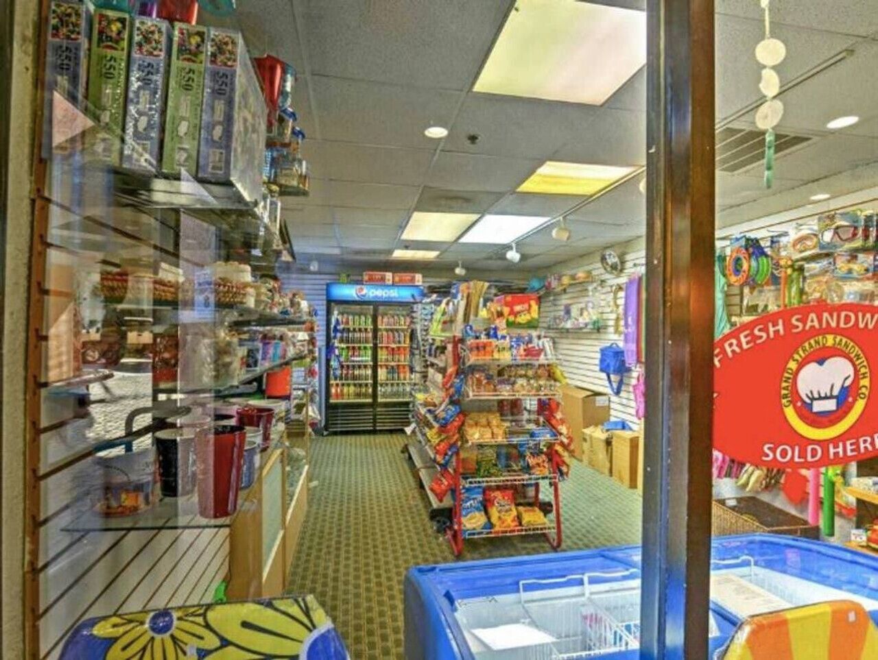 A store interior with shelves stocked with various products, a refrigerated section in the center, and a sign indicating fresh sandwiches sold here.
