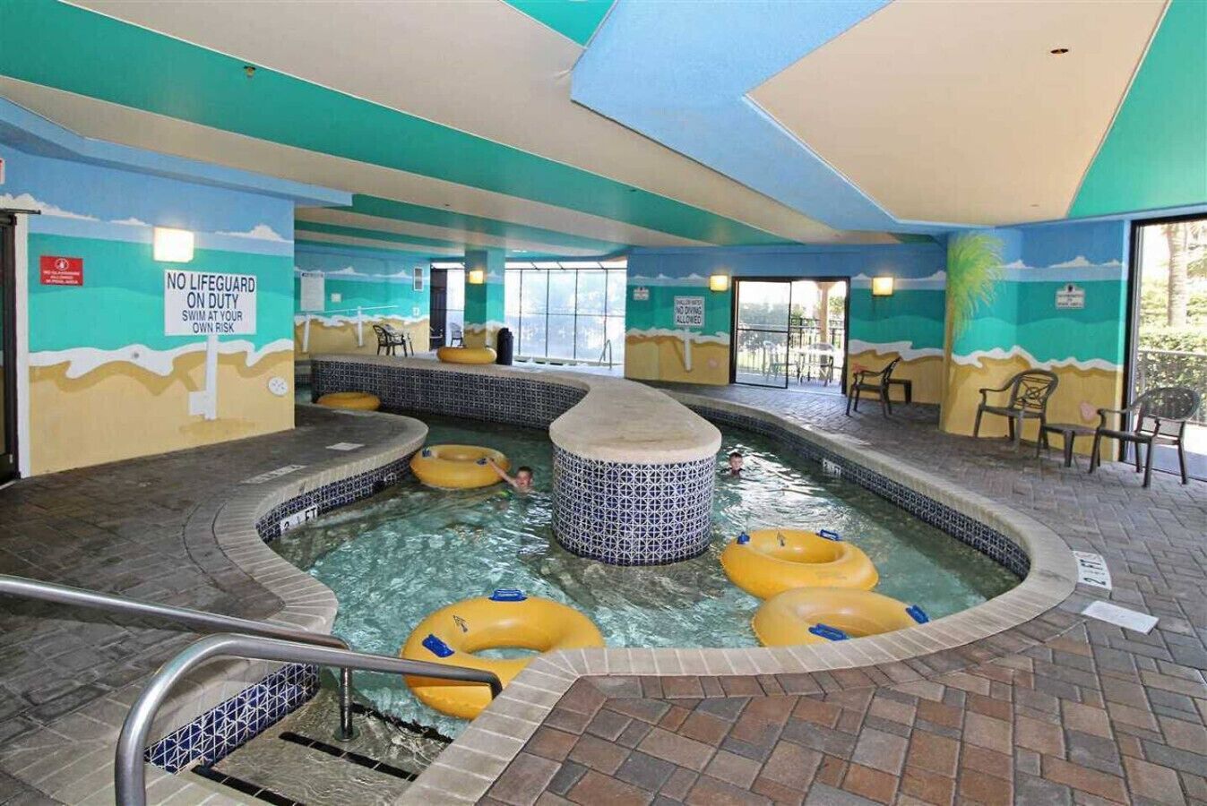 An indoor swimming pool with multiple hot tubs and inflatable rings, surrounded by a tiled floor and walls with a tropical mural.