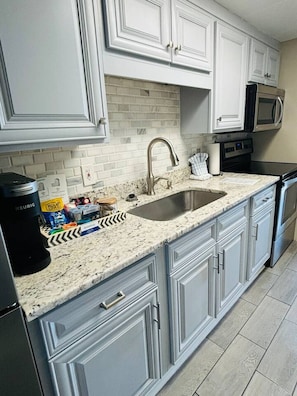 A kitchen with white cabinets, a granite countertop, a stainless steel sink, a microwave, and a coffee maker. - Beachfront Haven. Oceanfront (Myrtle Beach)